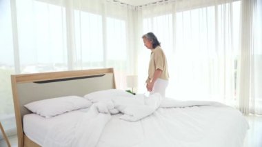 Yaşlı Asyalı kadın yatak örtülerini, battaniyeleri ve yatak örtülerini düzenleyerek ev işi yapıyor ve burnunu eliyle örterek alerjiye neden olabilecek toz ve kiri önlüyor..