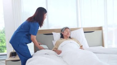 Kadın hemşire, kronik hastalık tedavisi gören yaşlı bir kadın hasta ile ilgileniyor. Onu nazikçe uyandırır ve, yatakta yastıkların karşısında yavaşça oturmasına yardım eder..