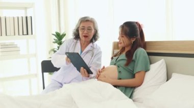 Kıdemli kadın doğum uzmanı hamile bir hastayla tanışıyor, tablet kullanarak hamilelik sağlığını tartışıyor, sağlık kayıtlarını tutuyor ve konforlu bir yatakta doğum öncesi tavsiyeler ve planlama sunuyor..