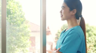 Mavi üniformalı Asyalı kadın doktor hastanede çalışırken kolları çapraz durarak kameraya gülümsüyor ve hemşirelik yapıyor..