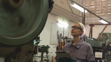 Güvenlik gözlüğü takan kadın torna operatörü metal ve güvenlik kulaklıkları gürültüsüne karşı koruyucu, parça üretim tesisinde ince metal levha damgalama makinelerini dikkatle kontrol ediyor..