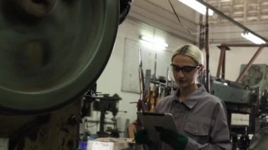Güvenlik gözlüğü takan kadın torna operatörü metal ve güvenlik kulaklıkları gürültüsüne karşı koruyucu, parça üretim tesisinde ince metal levha damgalama makinelerini dikkatle kontrol ediyor..