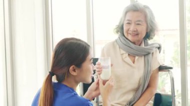 Yaşlı kadın tekerlekli sandalyeyle ilgilenen kadın hemşire, yaşlıların sağlıklı kalmasını sağlayan süt bardakları getiriyor ve osteoporoz ve D vitamini eksikliğini azaltıyor..