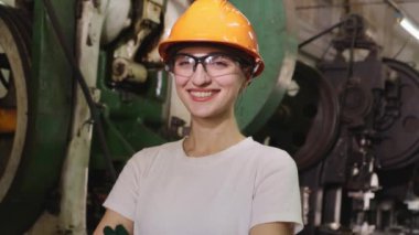Kadın fabrika teknisyeni, kasketli ve güvenlik gözlüklü oto yedek parça imalat fabrikasında çalışıyor. Çalışmadan önce makine güvenliğini kontrol ettikten sonra gülümsüyor..