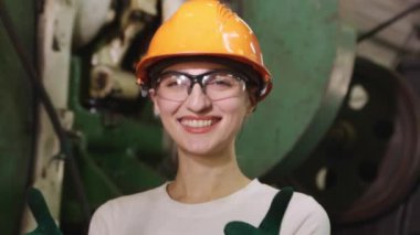 Kadın fabrika teknisyeni, kasketli ve güvenlik gözlüklü oto yedek parça imalat fabrikasında çalışıyor. Çalışmadan önce makine güvenliğini kontrol ettikten sonra gülümsüyor..