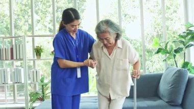 Kemik iltihabı olan yaşlı bir kadına bakan kadın hemşire, ayağa kalkmasına ve dikkatli yürümesine yardımcı olmak için bastona ihtiyaç duyar..