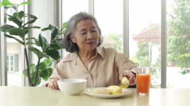 Sağlıklı, yaşlı, Asyalı bir kadın, C vitamini zengini, kolay sindirilebilen pirinç lapası, muz ve taze sıkılmış portakal suyuyla öğle yemeği yemek için oturur..