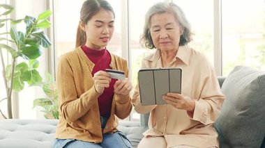 Kızı Asyalı yaşlı bir anneye tablet uygulamasından alışveriş yapmayı öğretiyor, ürünleri seçiyor ve kredi kartıyla kolayca ödüyor, her ikisi de alışverişi seviyor..