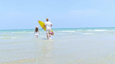 Mutlu yaz tatili seyahatleri: Baba ve kızı güneşli bir yaz gününde kumlu bir sahilde oynuyorlar, birlikte oynuyorlar ve şirin kıza sörf tahtası öğretmeyi..