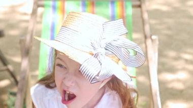 Şirin, uzun saçlı, şapkalı, kumlu sahilde bir plaj sandalyesinde oturan komik yüzünü gösteren mutlu bir kız. Denize gülümseyip kameraya bakıyor..