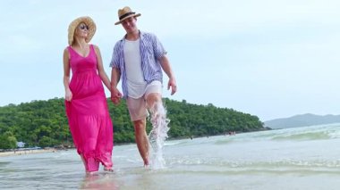 Beyaz kumlu sahilde ve mavi denizde el ele yürüyen mutlu çift: Çift yazlık elbiseler giyer, eş renkli elbiseler giyer ve koca sahilde yürürken günlük giysiler giyer.