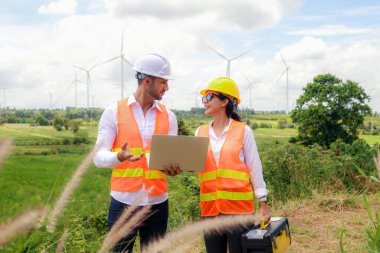 Erkek ve kadın mühendis, rüzgar çiftliği projesinde ellerinde dizüstü bilgisayar ve alet çantasıyla yürür ve tartışır. Bakım kontrolü, yüksek rüzgar türbini kulesi ve hedefe işaret eder..