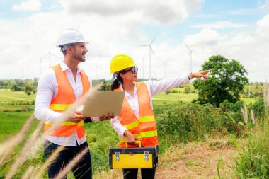Erkek ve kadın mühendis, rüzgar çiftliği projesinde ellerinde dizüstü bilgisayar ve alet çantasıyla yürür ve tartışır. Bakım kontrolü, yüksek rüzgar türbini kulesi ve hedefe işaret eder..