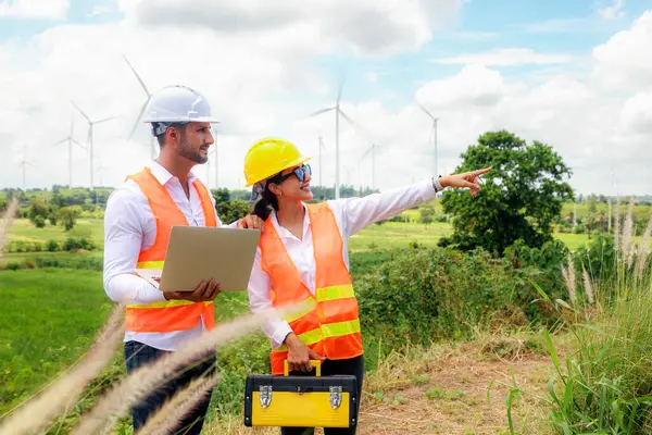Erkek ve kadın mühendis, rüzgar çiftliği projesinde ellerinde dizüstü bilgisayar ve alet çantasıyla yürür ve tartışır. Bakım kontrolü, yüksek rüzgar türbini kulesi ve hedefe işaret eder..