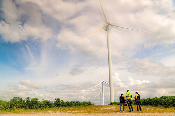 Команда инженеров мужского и женского пола вместе ходят по ветряной электростанции стоя на фоне облачного неба, делясь идеями о возобновляемых источниках энергии с Земли, рассматривая планы и собирая данные о завершенной миссии.