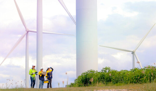 Команда инженеров мужского и женского пола вместе ходят по ветряной электростанции стоя на фоне облачного неба, делясь идеями о возобновляемых источниках энергии с Земли, рассматривая планы и собирая данные о завершенной миссии.