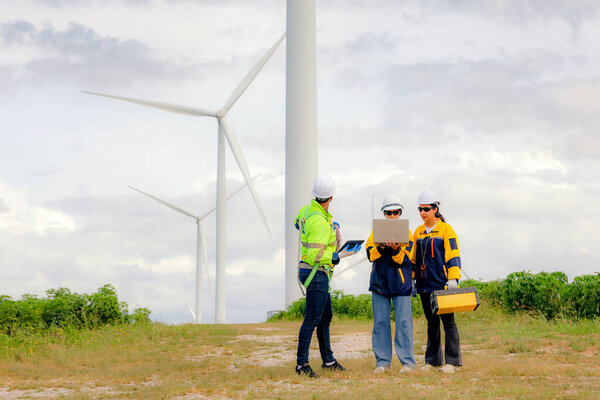 Команда инженеров мужского и женского пола вместе ходят по ветряной электростанции стоя на фоне облачного неба, делясь идеями о возобновляемых источниках энергии с Земли, рассматривая планы и собирая данные о завершенной миссии.