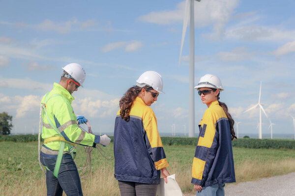 Команда инженеров мужского и женского пола вместе ходят по ветряной электростанции стоя на фоне облачного неба, делясь идеями о возобновляемых источниках энергии с Земли, рассматривая планы и собирая данные о завершенной миссии.