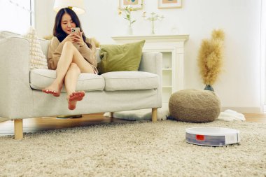 Genç Asyalı kadın elektrikli süpürge, temiz halı ve ahşap döşeme kullanarak ev işi yapıyor. Kanepede oturmuş akıllı telefonla oynarken gülümsüyor ve ev işi yapmaktan yorulmuyor..