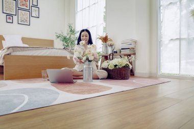 Asyalı bir kadın tatilini evinde, yatak odasında geçiriyor. Evini güzelce süslemek için vazoda pastel renkli plastik güller düzenliyor..