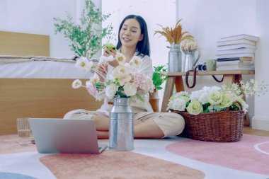 Asyalı bir kadın tatilini evinde, yatak odasında geçiriyor. Evini güzelce süslemek için vazoda pastel renkli plastik güller düzenliyor..