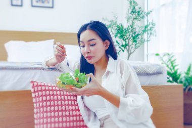 İnce ve ince bir kadın vejetaryen sebzeli yeşil salata yiyor. Genç Asyalı bir kadın, yatak odasında rahat bir şekilde oturmuş organik sebze yiyor ve şekersiz ve yağlı tatlandırıcı kullanmıyor..