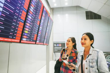 Uluslararası bir yolculuk için biletleri olan iki Asyalı kadın yolcu kalkış terminalinde durdu ve kalkış panosuna kendi uçuşlarını kontrol etmek için ilgiyle baktı..