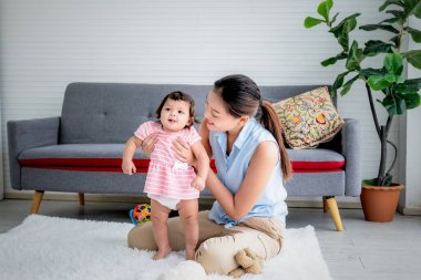Asyalı anne gülümseyerek ve mutlu bir şekilde 7 aylık kızının Asya aile ve çocuk gelişimi kavramında ayakta durmayı öğrenmesini destekliyor..