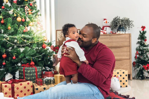 African family, Nigerian father kissing 4-month-old baby newborn son, with smiling and happy together,  blur background of Christmas tree and gift boxes. to African family on holiday and baby infant concept.
