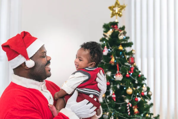 African family, 4-month-old Nigerian baby newborn son and father is wearing a red Santa costume, smiling and happy together, with blur background of Christmas tree, to Christmas and baby concept.