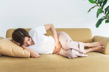 Kanepede yatan Asyalı çekici bir kadın hasta ve karnı ağrıyor ya da kramp geçiriyor, her kadının hastalığı ve sağlık konsepti....