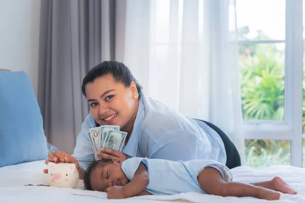 Baby newborn son half-Thai, half-Nigerian, 2 months old is sleeping on a white bed and mother is putting money in a piggy bank, concept is to saving money for the future of the baby.
