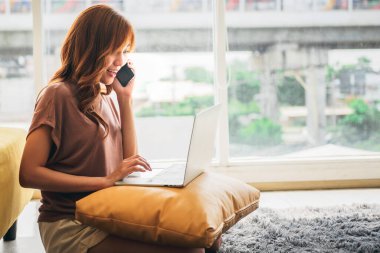 Asian woman, attractive and young, sitting on floor talking a mobile phones and using a notebook computer with smile and happy, to people, work from home and shopping online concept.