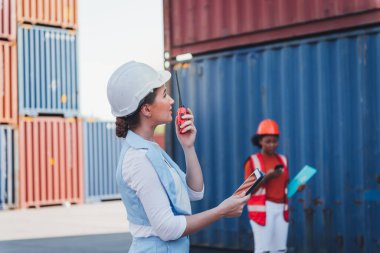 Beyaz kadın mühendis iletişim radyosu kullanıyor ve konteynır endüstrisinde çalışıyor, lojistik ulaşım ithalat-ihracat kargosunda, iş taşımacılığı ve depolama terminali kavramına.