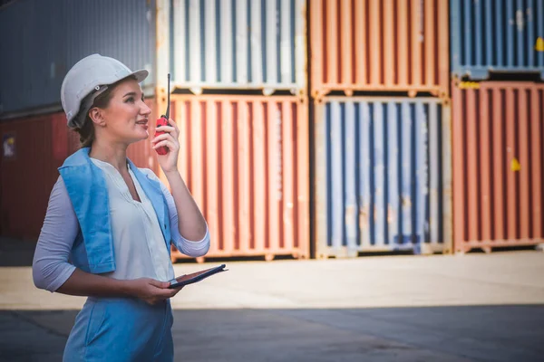 Caucasian female engineer using a communication radio and working in containers industry, logistic transport Import-Export Cargo, to business shipping and storage port terminal concept.