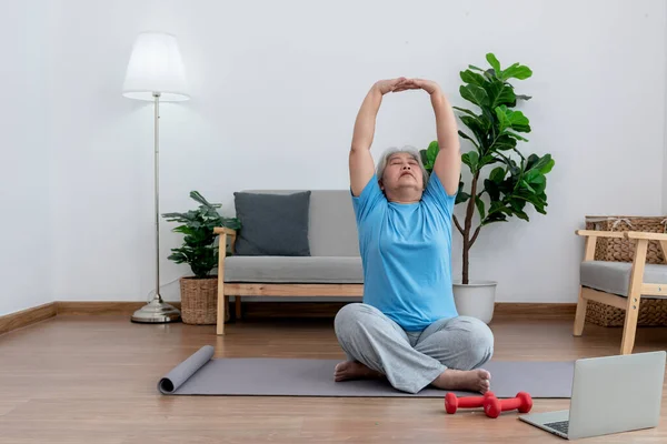 Asian elderly women are practicing yoga in the basic position in her living room, which is a warm-up and meditation exercise, to elderly health care concept.