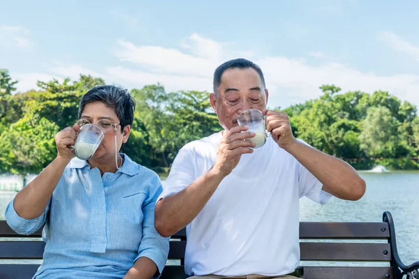 Asian elderly couple sitting, holding glass and drinking milk, they smile and happy together in the the park. to drink milk for good health of retirement age concept.