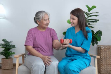 Yaşlı bir Asyalı kadın bir hemşire tarafından denetlenirken fizik tedavi görüyor. Oturma odasındaki kanepede hastaya egzersiz yaptırmak için halterleri kullanarak oturmak. Sağlık hizmeti kavramı