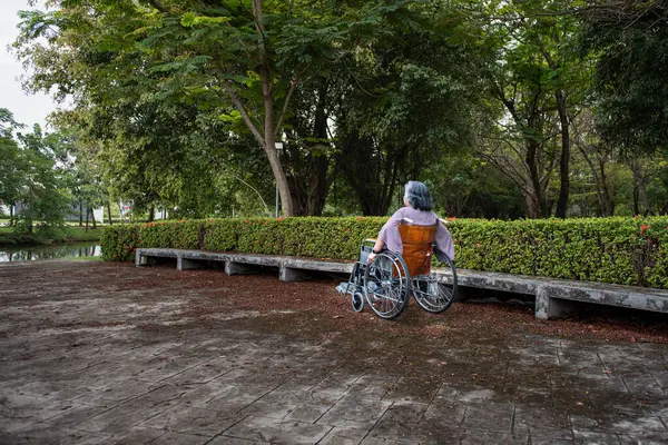 Elderly Asian female patient sitting in a wheelchair Using hands to move the wheel of a wheelchair forward in the park. Elderly people with heart disease and health care concept.