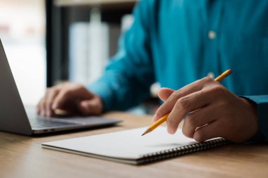 Bir adam kalemle deftere yazı yazıyor. O, işine odaklanmış ve ciddi bir ruh hali içinde. Defter ahşap bir masanın üzerine yerleştirilmiş ve yakınlarda bir laptop var.