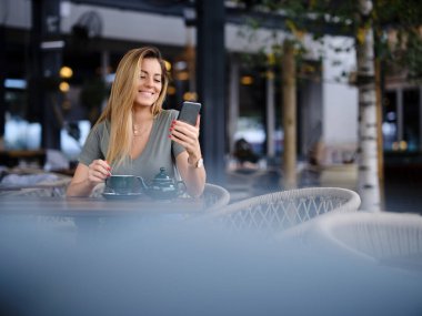 Bir kadın kafeteryada oturmuş, çaydan zevk alıyor ve telefonunu internette sohbet için kullanıyor..