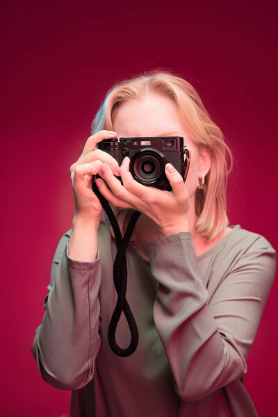 A happy blonde photographer is holding camera while standing in front of the magenta background. Viva magenta.