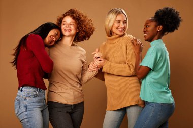 Four multiracial female friends hugging and laughing in studio isolated on brown background.