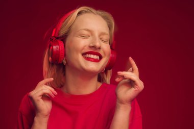 Kırmızı arka planda müzik dinleyen mutlu bir kız kablosuz kulaklıklarla ve gözleri kapalı gülümseyen..