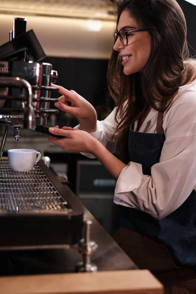 A happy waitress making fresh coffee on a espresso machine in cafeteria.