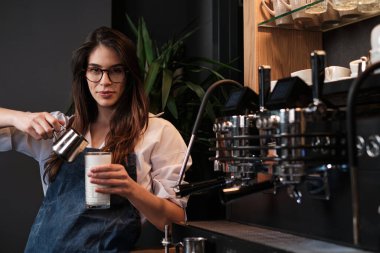 Kahve makinesinin yanında duran bardağa taze kahve hazırlayan ve süt ekleyen mutlu bir barista..