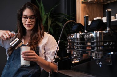 Kahve makinesinin yanında duran bardağa taze kahve hazırlayan ve süt ekleyen mutlu bir barista..