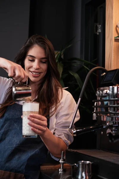 stock image A happy barista preparing fresh coffee and adding milk into a glass while standing next to a coffee machine.