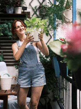 Tulumlu, kızıl saçlı, neşeli bir yüzü olan bir kadın balkon bahçesinde saksı çiçeği tutuyor.