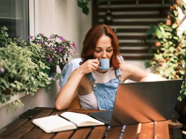 Kızıl saçlı kadın, güneşli bir balkonda çiçekli laptopunu kullanırken bir fincan kahveden hoşlanıyor.